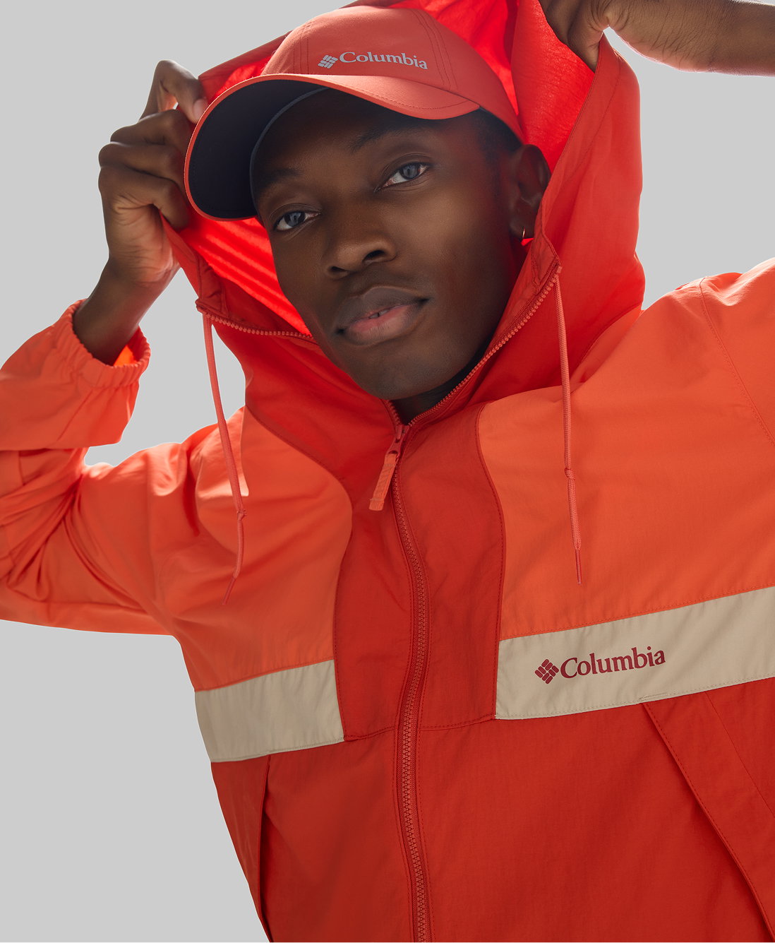 A man wearing a red windbreaker with the hood up, as well as a red ball cap,against a grey background.