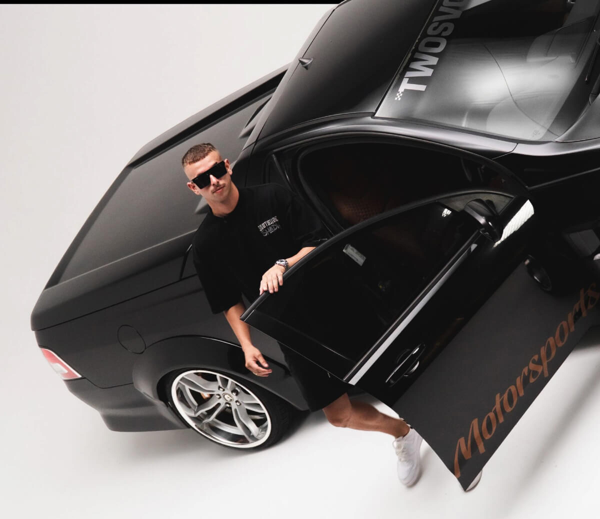 Person wearing black Two Svge sunglasses standing next to black sports car in minimalist white studio