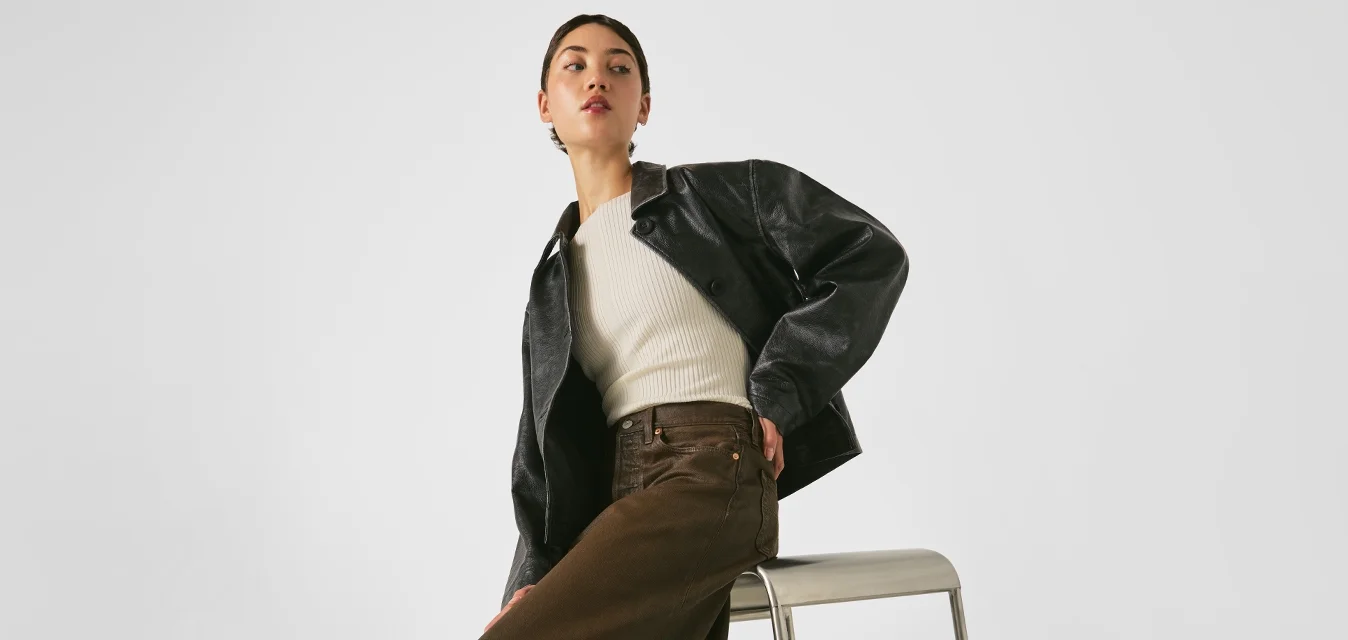 Frau, die auf einem silbernen Metallhocker sitzt, trägt eine schwarze Jacke, ein cremfarbenes Oberteil, einen braunen Rock, weiße Socken und beige Absätze vor einem schlichten Hintergrund.