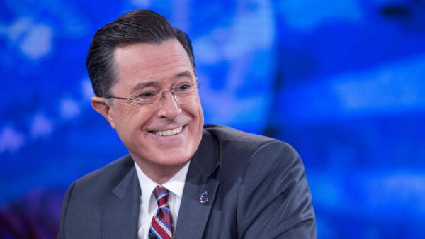 Stephen Colbert, wearing a blue suit and a striped blue, red and white tie, smiles off to the side. He is pictured from the shoulders up. 