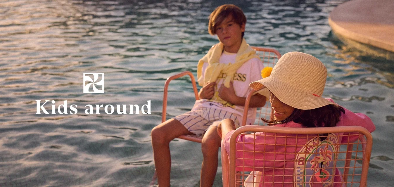 Deux enfants se détendent sur des chaises, les pieds dans l'eau peu profonde, l'un portant un grand chapeau de soleil et un sweat-shirt rose, l'autre en short et chemise blanche.