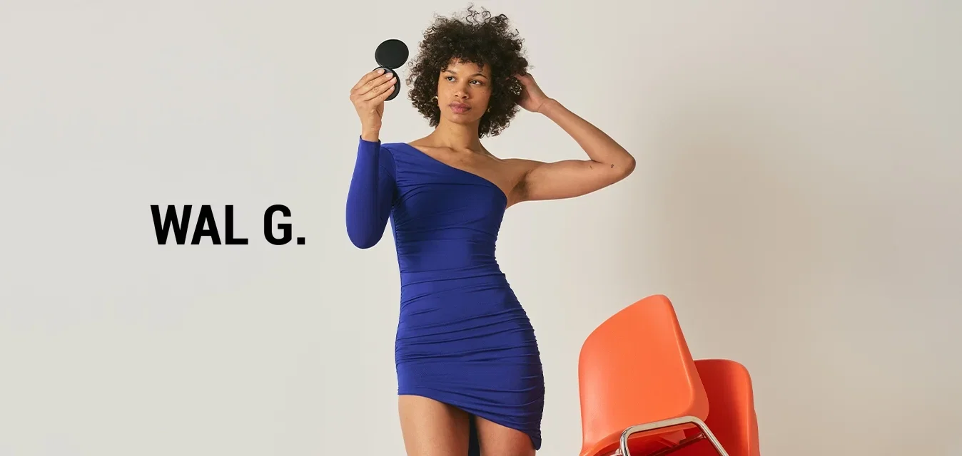 Femme en robe bleue asymétrique tenant un miroir compact rond, debout à côté d'une pile de quatre chaises orange sur un fond uni.