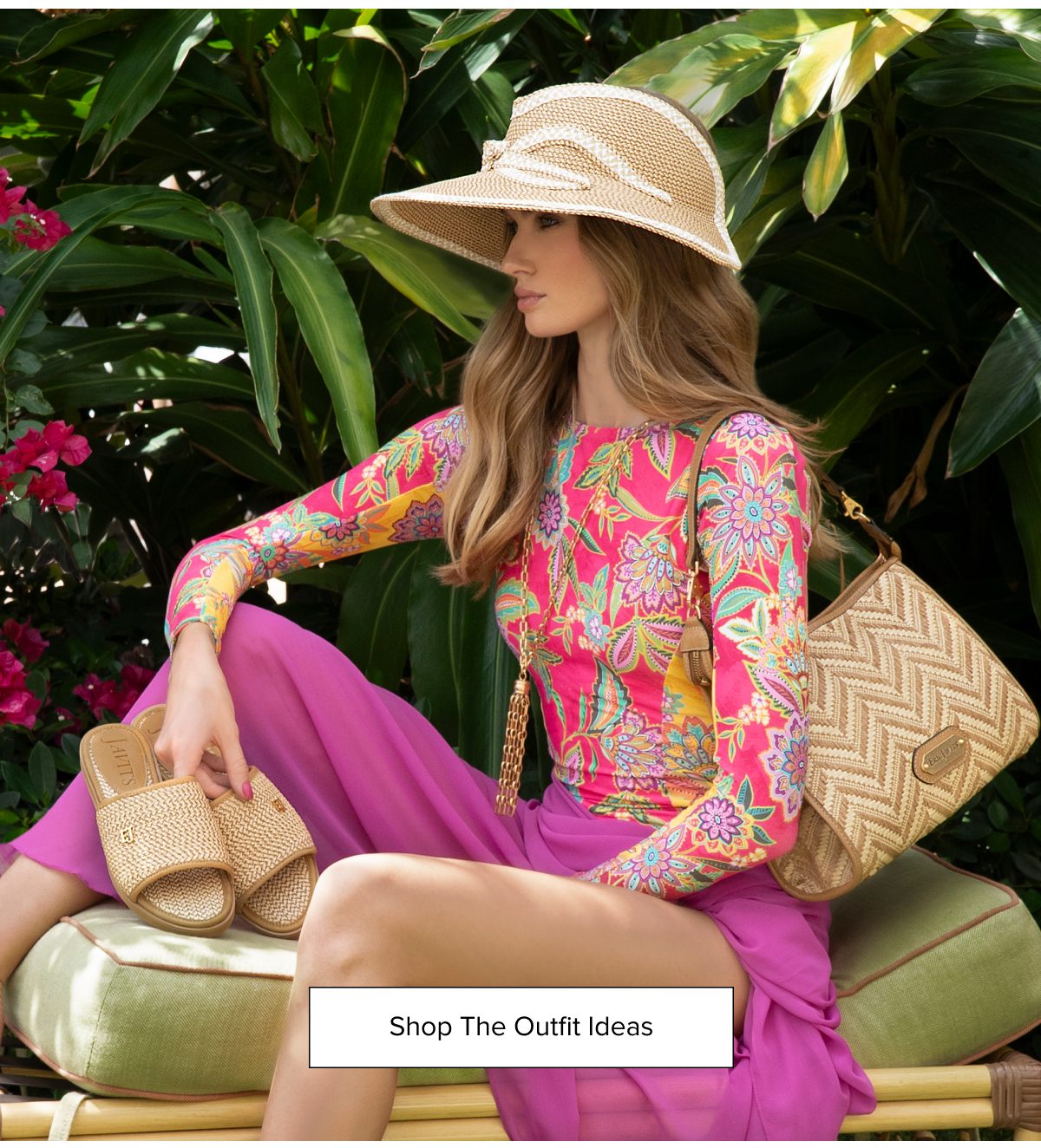 Woman in colorful outfit and hat sitting among tropical plants.