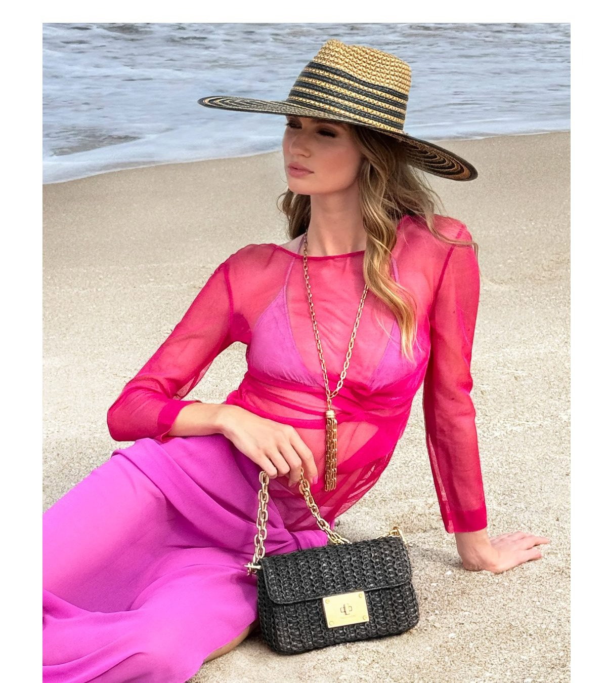 Woman in a pink outfit and straw hat sitting on the beach with a black handbag.