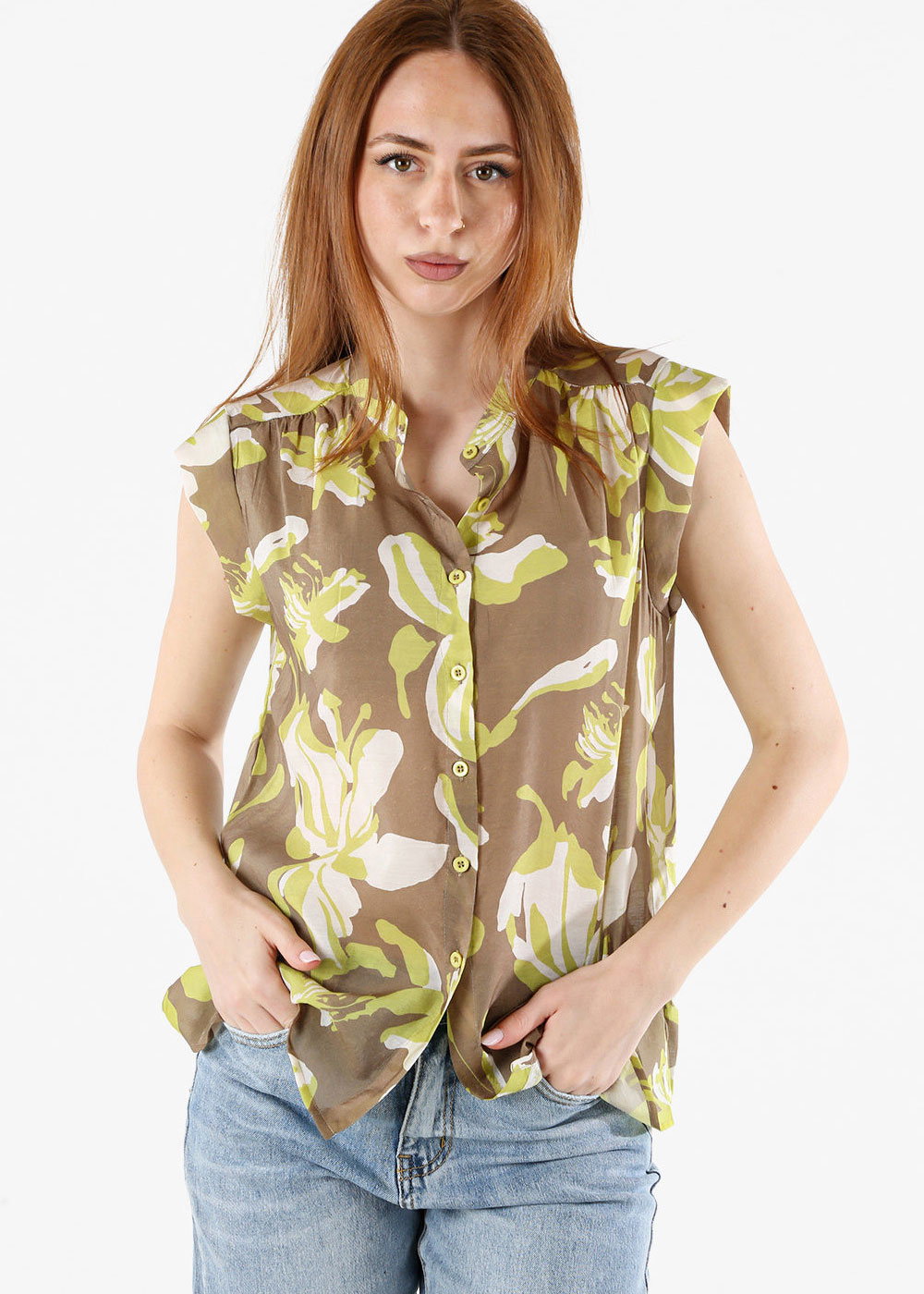 Woman wearing sleeveless brown and lime floral button-up blouse and blue jeans, hands in pockets, neutral expression