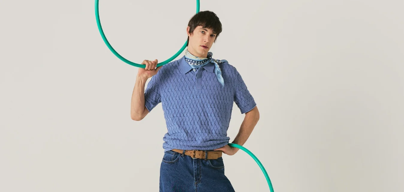 Persona con suéter azul y jeans posa con un aro de hula verde, de pie contra un fondo claro y liso.