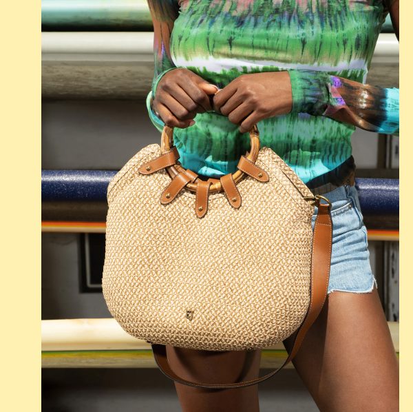 A person holding a woven straw handbag with leather straps, wearing a colorful tie-dye top and denim shorts.