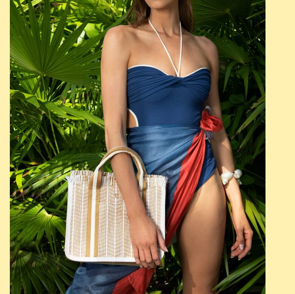 A woman in a blue swimsuit with a red wrap and a straw bag, surrounded by tropical plants.