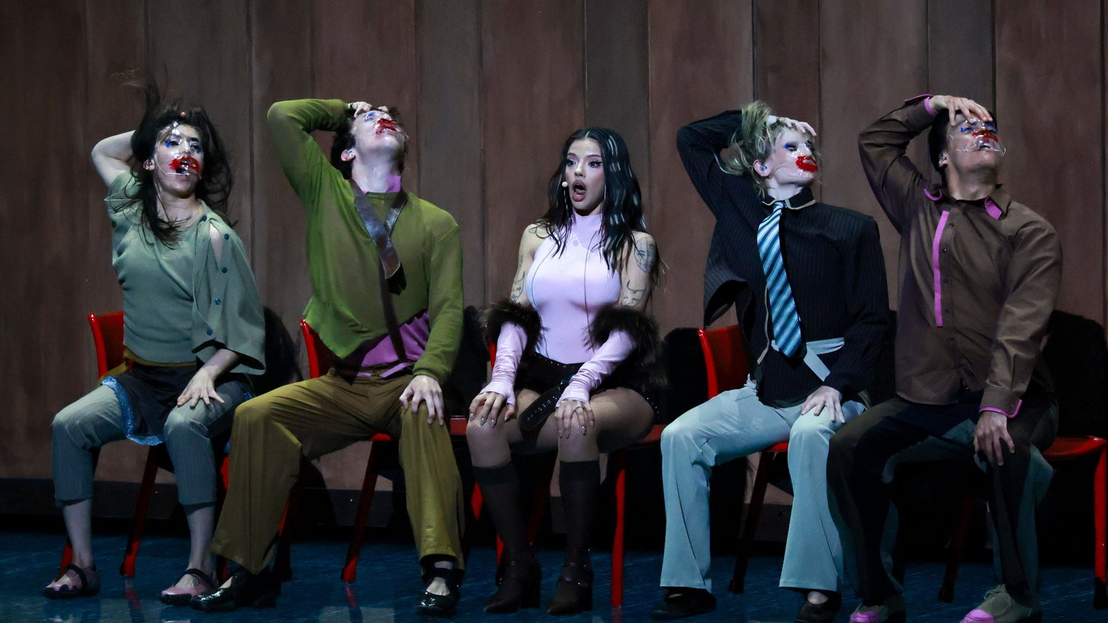 Dara performs at Eurovision. She sits in the middle of a group of 5 people sat on a bench. They each have one hand on their head looking to their left, except Dara, who has two hands on her knees and looks shocked.