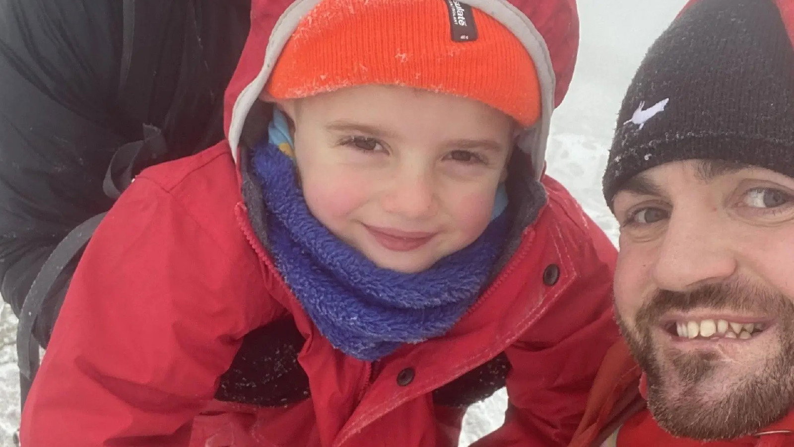 A four-year-old boy wearing a red coat, orange wool hat and a blue scarf smiles in the snowy weather next to his Dad, who wears a black wool hat.