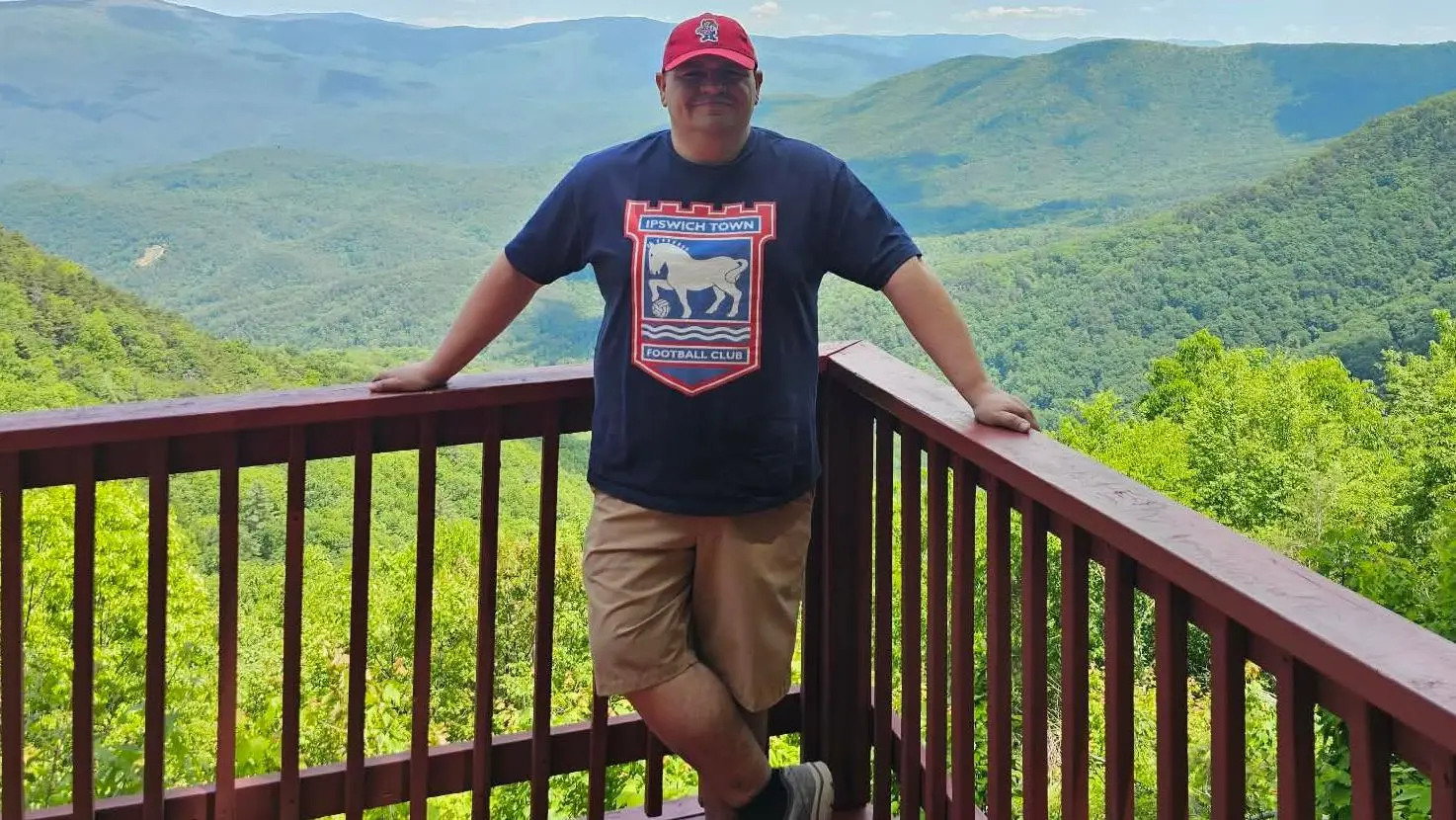 Zach Peterson stands on a balcony, with a large expanse of lush green trees and hills in the distance behind him. He wears a red cap and a navy blue T-shirt with the Ipswich Town FC embelem on it.
