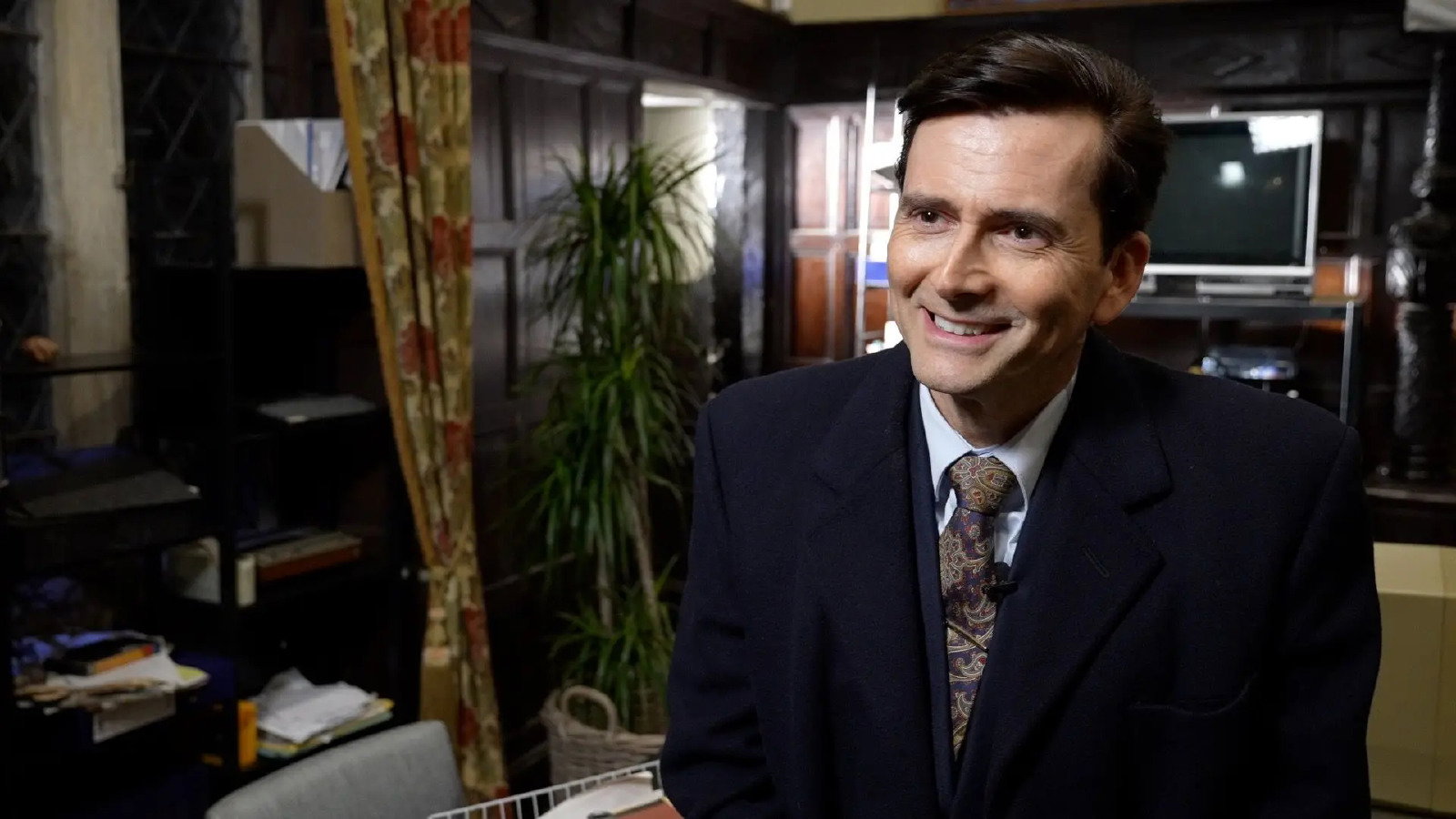 David Tennant smiles as he is interviewed on the TV set of Rivals. He wears a black suit jacket, a pale blue shirt and a brown paisley tie. The room he is in is cluttered, with a curtain, houseplant and TV in the background.