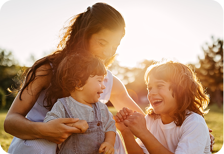Mutter sitzt mit zwei Kindern im Freien, alle lachen und spielen miteinander in der Sonne.