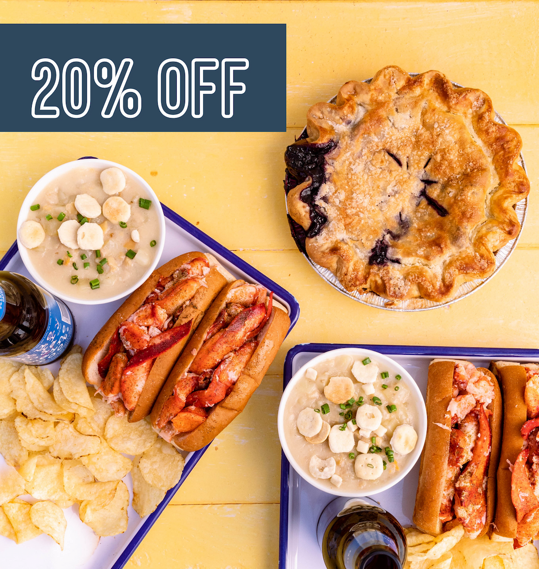 Two trats of lobster rolls, chips, and blueberry sodas on a yellow picnic table with a large blueberry pie next to them.