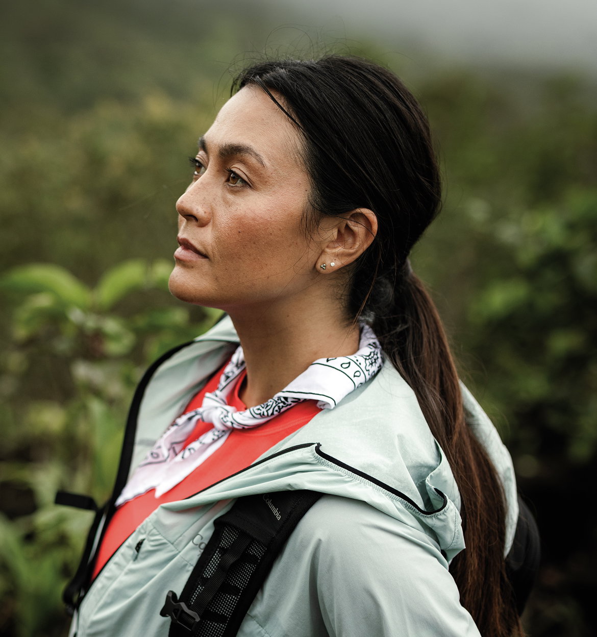 A woman wearing red shirt, blue windbreaker, and purple neckerchief against a lush jungle background.