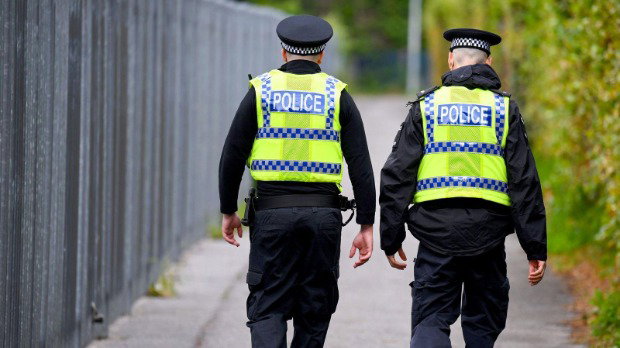 Two police officers are walking down a street. They are both wearing hi-vis vests over a black uniform and wearing black hats. There are bushes to their right and a metal fence to their left.
