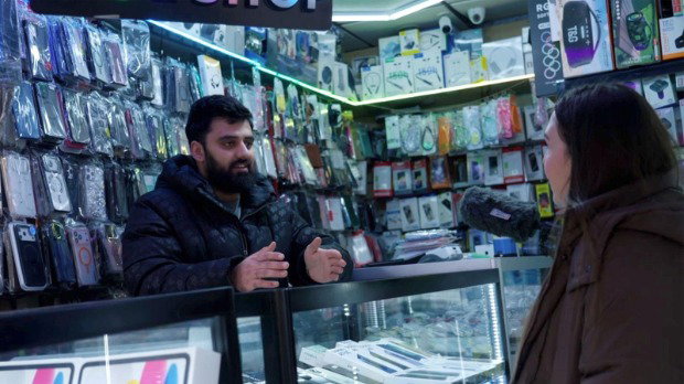 Shop worker is standing behind a glass display counter in a mobile phone accessories shop, speaking to a reporter holding a microphone, with walls lined with packaged phone cases and electronics.