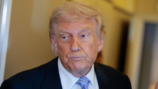Donald Trump is pictured close-up, looking to the side. He wears a black suit with a blue tie and is pictured inside a yellow hallway.