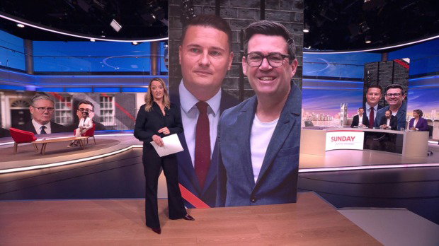 Laura Kuenssberg stands in front of a TV studio that has a panel showing pictures of Wes Streeting and Andy Burnham. In the background is Kemi Badenoch sat on a red chair to the left, and a panel of three people all sat behind a desk on the right.