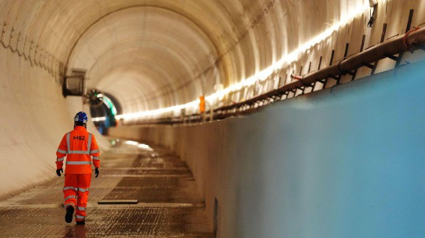 A man in orange hi-vis clothing, branded with the logo of HS2, walks through a well-lit tunnel.
