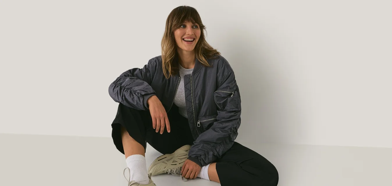 Jonge vrouw in casual kleding en sneakers zit op de vloer tegen een eenvoudige witte muur, glimlacht en kijkt opzij.