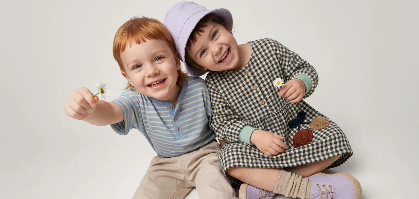 Twee glimlachende kinderen zitten op de grond, houden kleine bloemen vast, de een draagt een paarse hoed en een geruite jurk, de ander een gestreept hemd en broek.