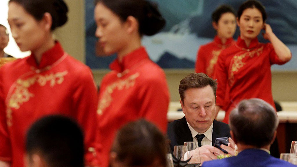Elon Musk sits at a table looking at a smartphone he holds, while women in tradition red Chinese dresses walk around tables. The heads of other guests at a dinner are blurred in shot.