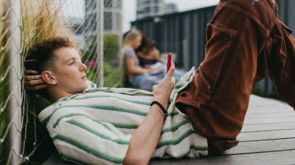 A young man is lying down on a bench holding and scrolling on his smartphone.