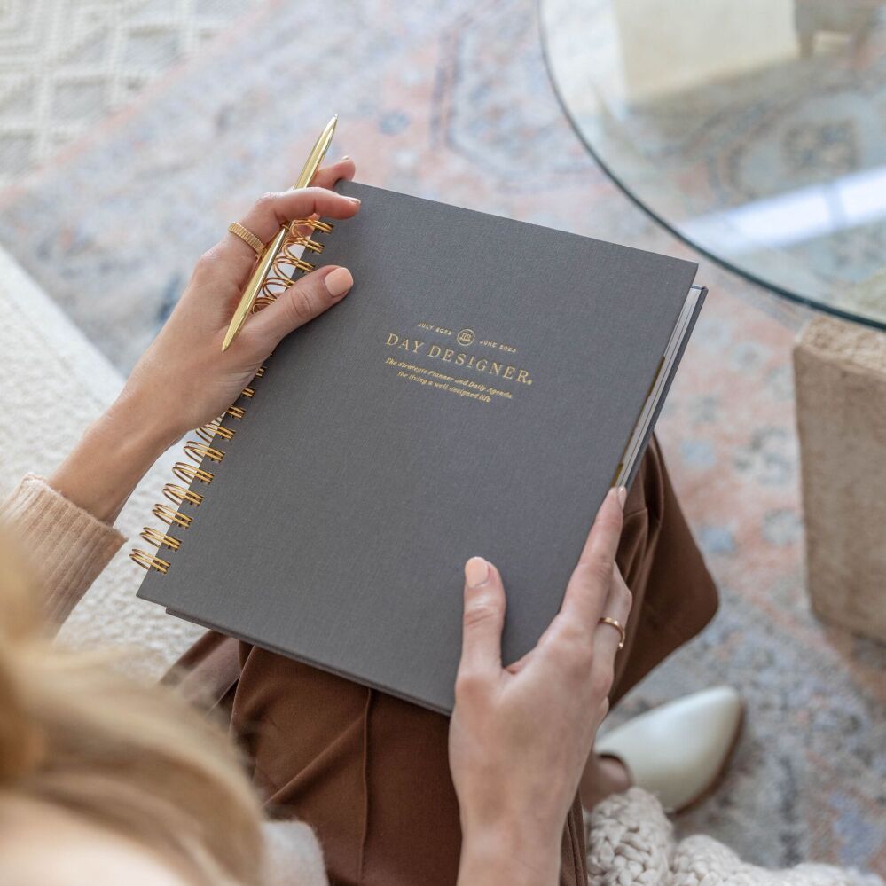 hands holding charcoal gray day designer planner, glass table, rug