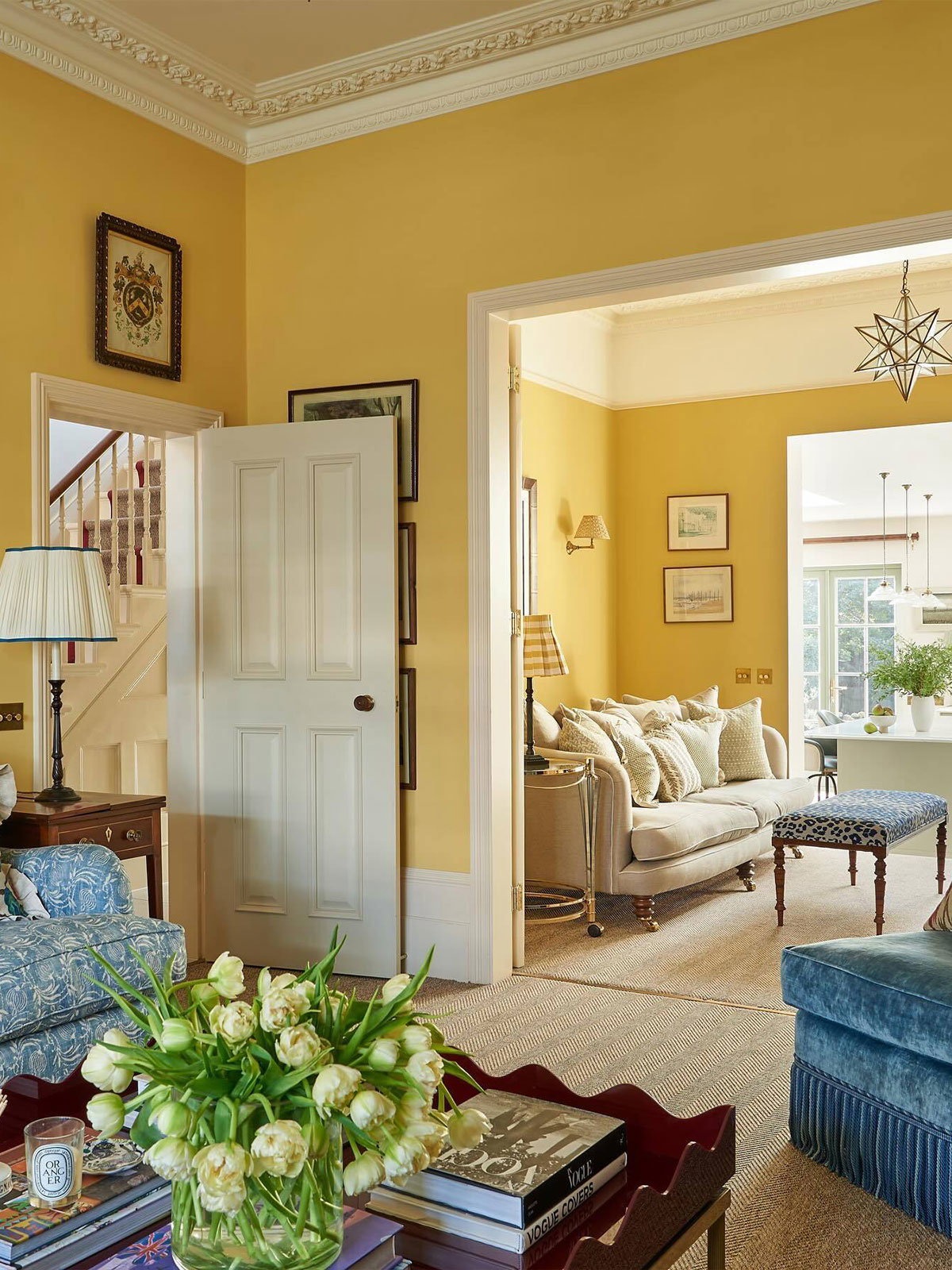 Sunlit living room with yellow walls, blue and beige furniture, and a vase of pale tulips on the coffee table.