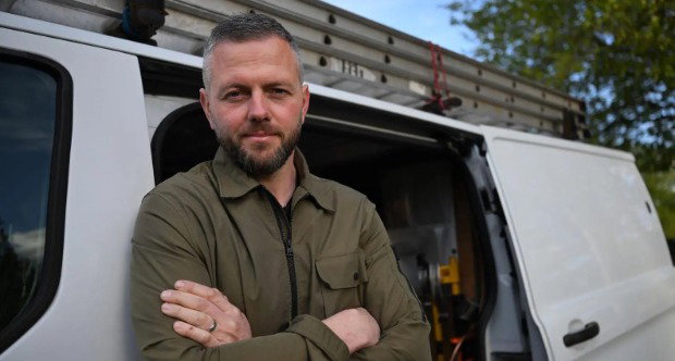 Robert Kenyon, a man with short grey hair and a beard, wears green overalls and leans against a white van with its door open with his arms crossed over his chest.