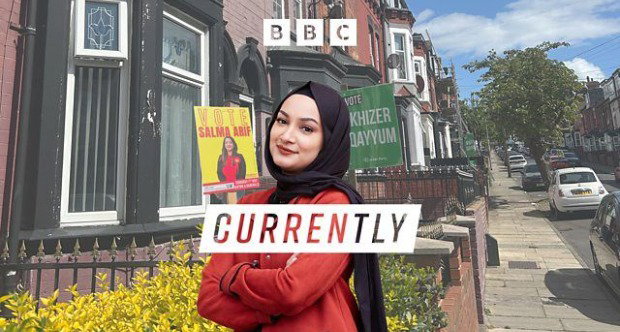 A woman dressed in red and wearing a black headscarf stands with her arms folded down a residential street. Behind her are houses and cars, as well as two signs endorsing local election candidates.