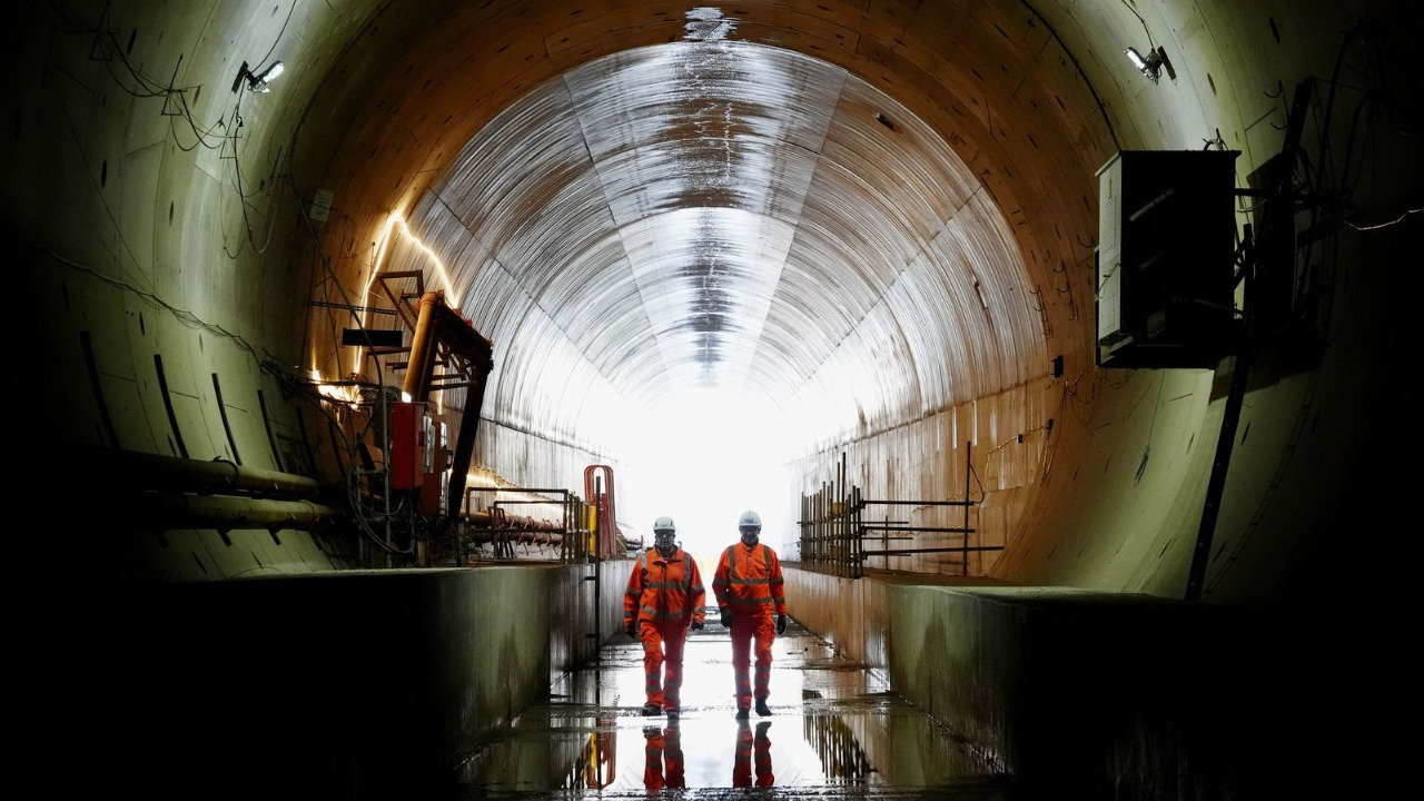 Two people in orange overalls and white hard hats walk along a large round tunnel. There’s machinery surrounding them, including lighting and wires, and their reflections show in the shiny floor surface.
