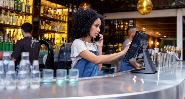 A young woman speaks on the phone while inputting information into a point-of-sale terminal in a busy bar, with glasses on the bar next to the worker and other bar tenders in the background next to bottles on display on the shelves.