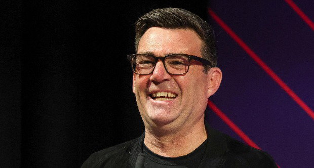 Andy Burnham, who has short dark hair and wears glasses, smiles as he speaks into a small black microphone. He stands in front of a dark purple background with diagonal red stripes running through it.