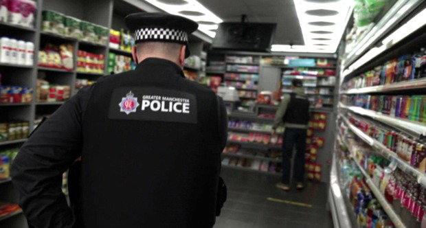 A Greater Manchester Police officer wearing a black police uniform enters a corner shop with shelves of soft drinks and grocery products. There is a man with his backed turned standing by the till.