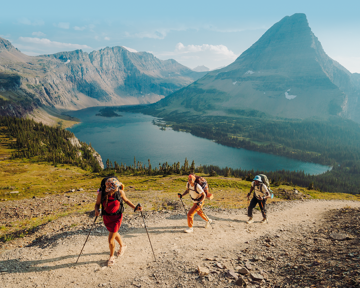 An image of three hikers ascending a rocky trail in a mountainous landscape with a large lake and rugged peaks in the background.