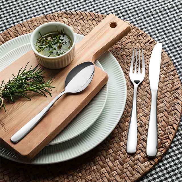 A table setting featuring a white fluted plate on a woven placemat, topped with a small wooden board. On the board are a spoon, fresh rosemary sprigs, and a dish of herb-infused oil. Beside the plate are a knife and fork. The backdrop is a black and 