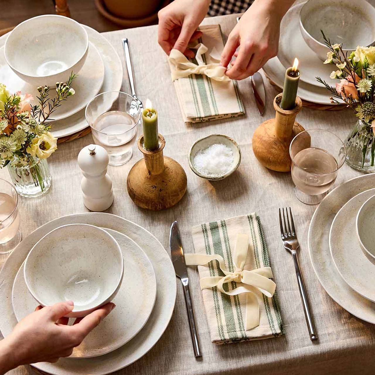 A neatly set dining table with plates, bowls, glasses, and cutlery. There are green candles in holders, small bouquets of flowers, and a salt dish. Napkins tied with ribbon are placed near each setting. Two people are arranging dishes. The tablecloth