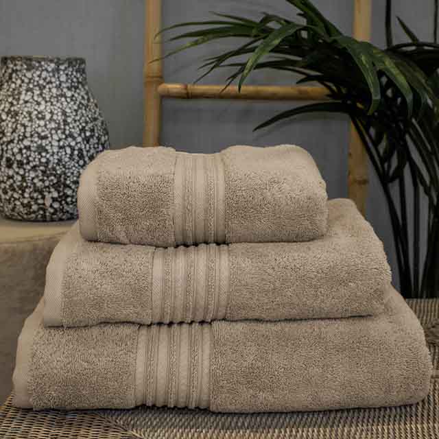 Stack of three beige towels, neatly arranged, with a decorative ceramic pot featuring a speckled pattern in the background. A bamboo ladder and green plant add a natural touch.