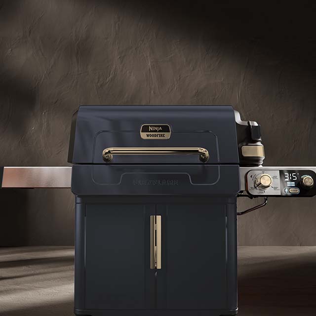 A black and silver Ninja Woodfire outdoor grill against a dark background. The grill features a main cooking chamber with a handle, a digital control panel displaying '315', and a side shelf.