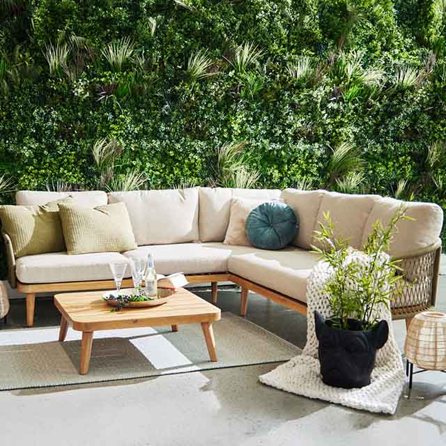 A cozy outdoor seating area with a beige corner sofa, pillows, a wooden table, potted plants, and a lush green vertical garden.