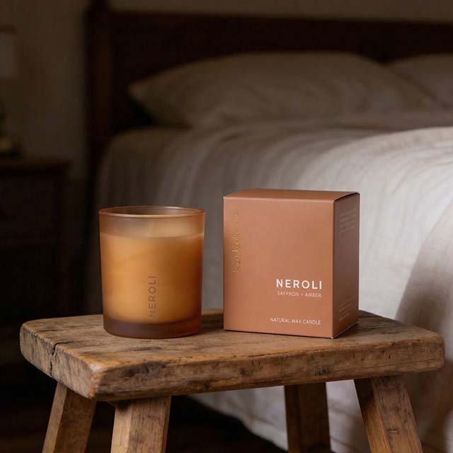 A frosted orange candle and its box, labelled 'Neroli, Saffron, Amber,' rest on a rustic wooden stool in a cosy bedroom. A neatly made bed is visible in the background.