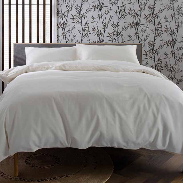 A neatly made bed with white pillows and a duvet, set against a wall with a floral bamboo pattern. The floor is wooden, and there's subtle natural light coming through a window with vertical wooden slats.