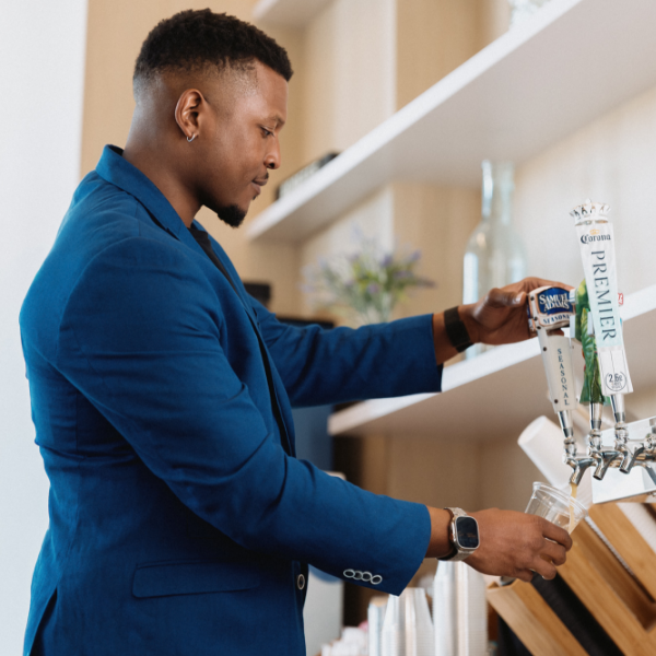 A man in a blue blazer pours draft beer from a tap into a clear plastic cup. He stands at a modern bar counter with shelves and decorative items in the background. The beer taps include clearly visible handles labeled "Samuel Adams" and "Corona Premier." The scene feels casual and contemporary, with neutral tones and soft lighting. The focus is on the pouring action and the bar environment.