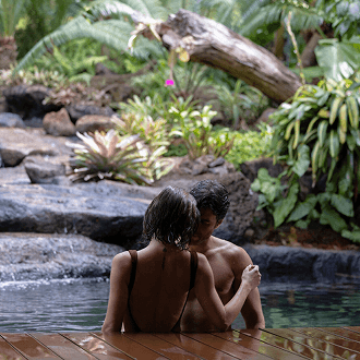 A man and woman sit side-by-side in a shallow pool beside natural rock and greenery.