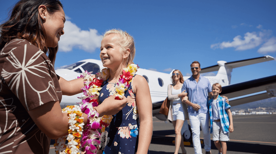 A staff member places a flower lei on a girl beside a private jet, as her parents and brother walk up behind them.