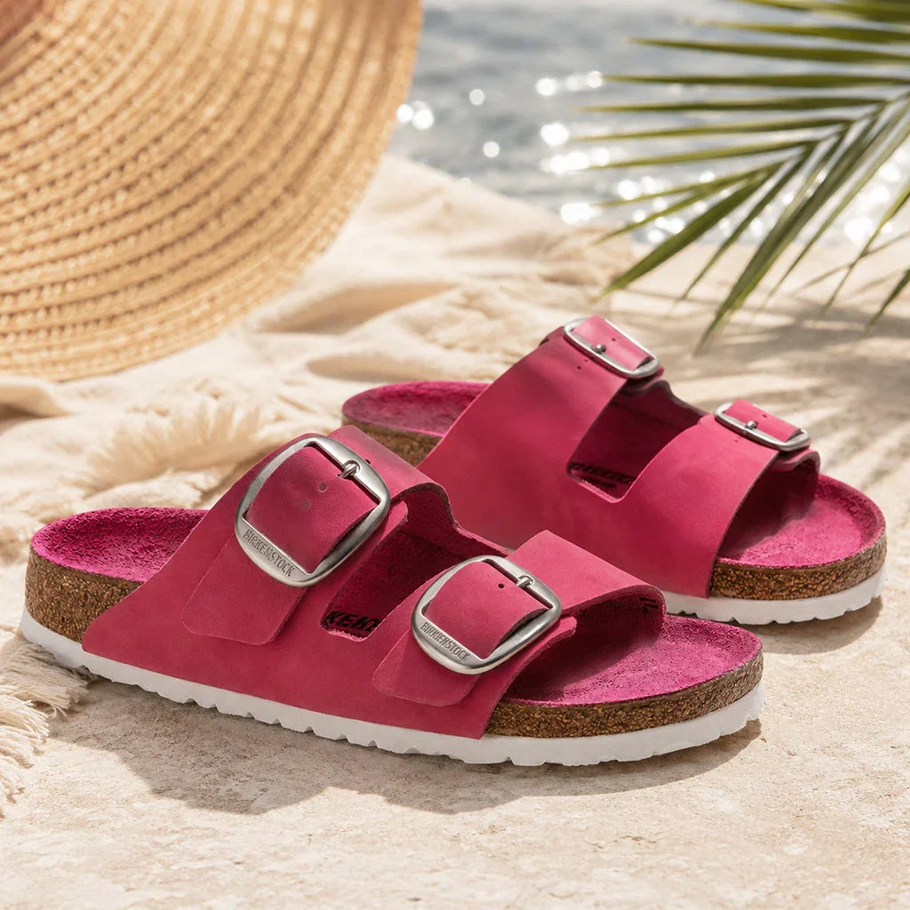 Two pink Birkenstock sandals displayed on sandy beach with straw hat and palm fronds in background.