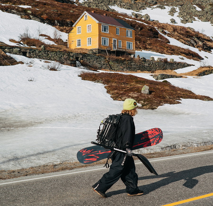 Burton rider, Ylfa walking on the road with a yellow house in the background surrounding by melting snow on a brown-colored mountain.