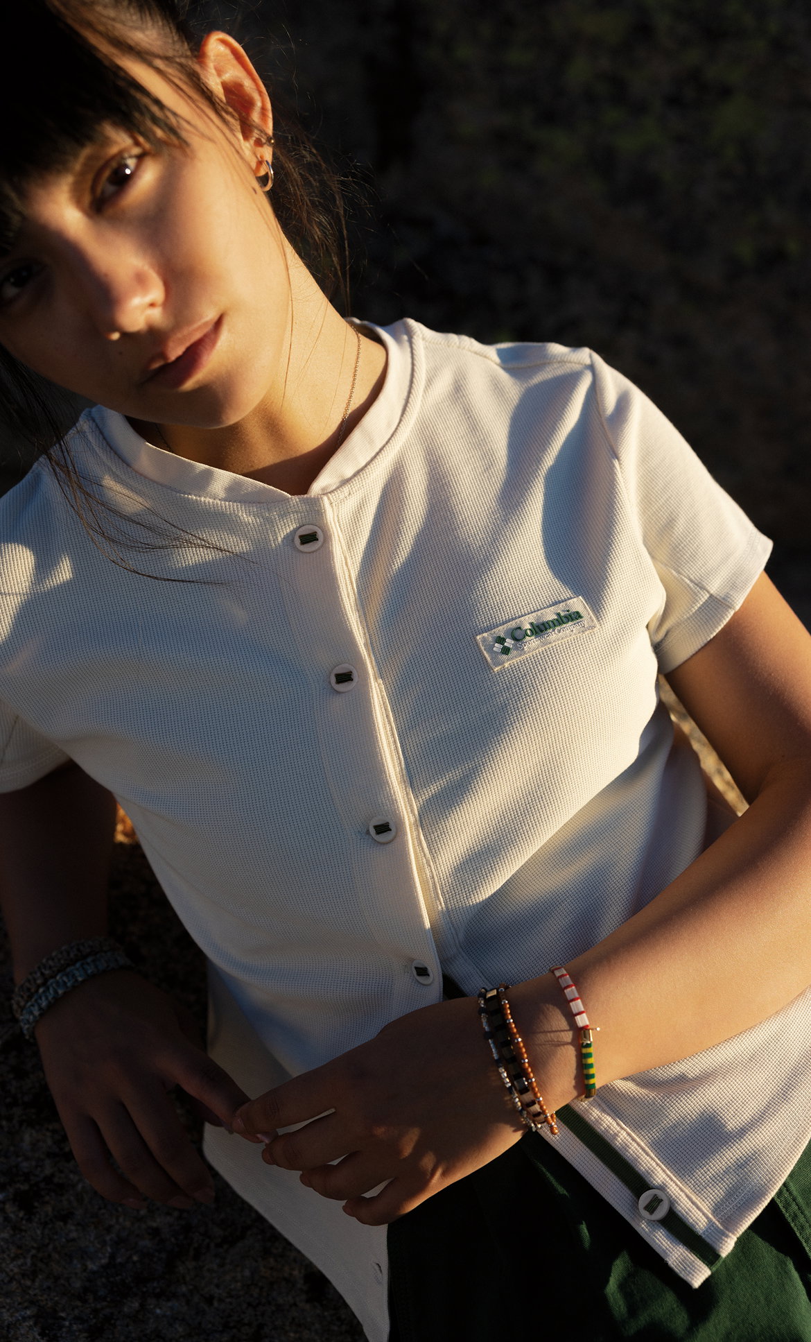 A woman wearing a white short sleeve button-up top, green shorts, and bracelets in the outdoors.